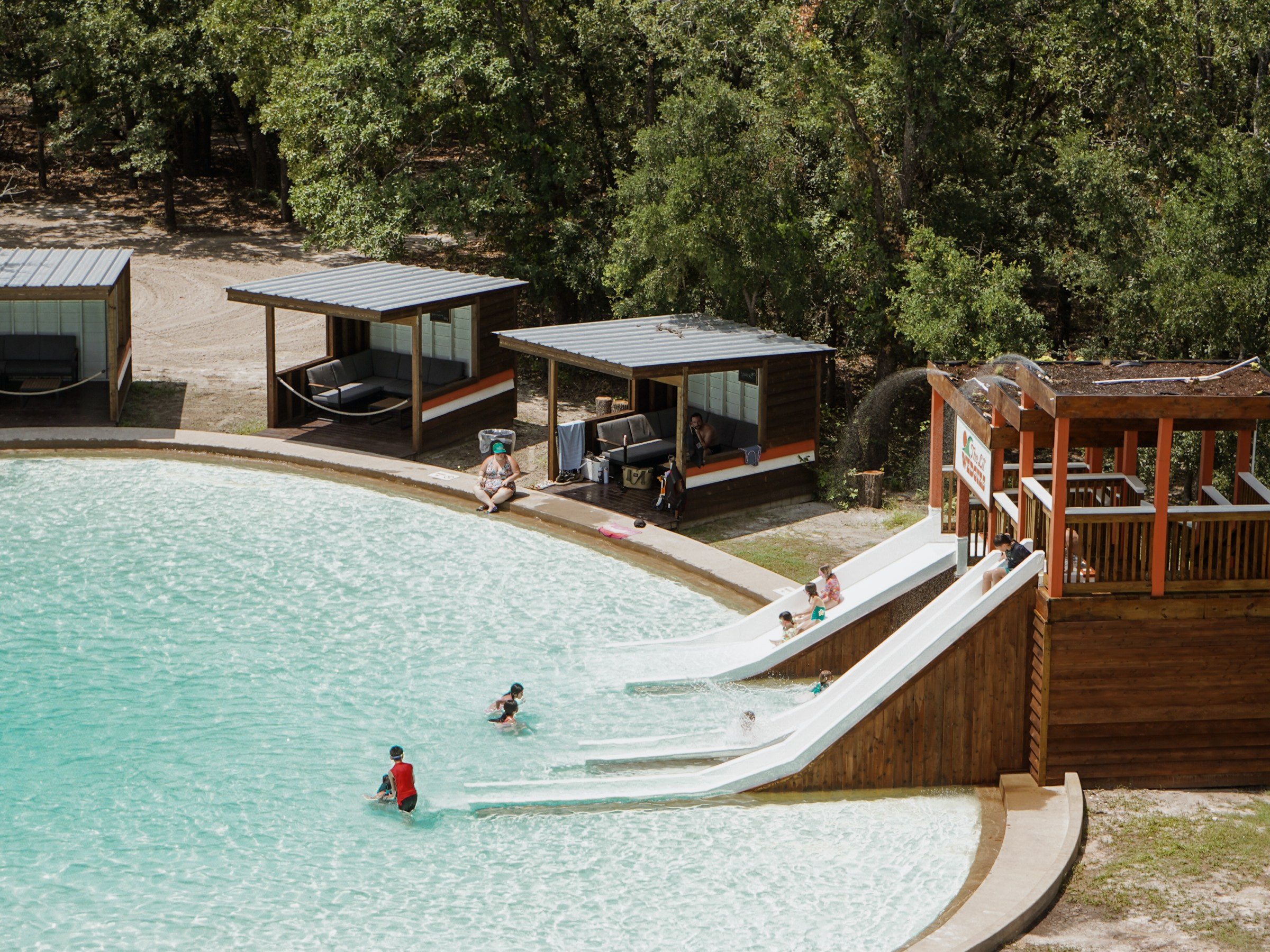 People enjoying a pool with slides and cabanas surrounded by trees.