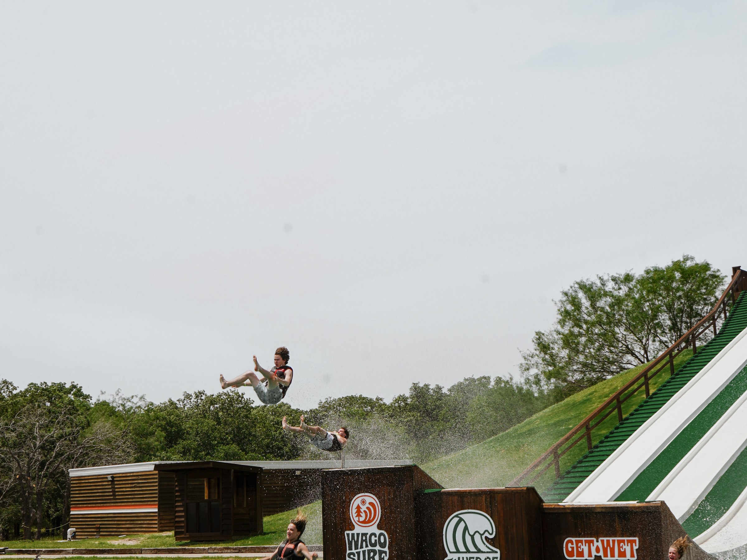 People flying off a large outdoor water slide into a pool, surrounded by trees.