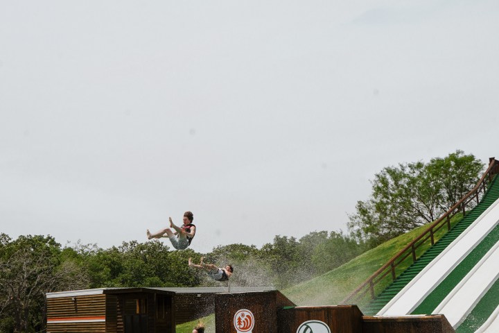 People flying off a large outdoor water slide into a pool, surrounded by trees.