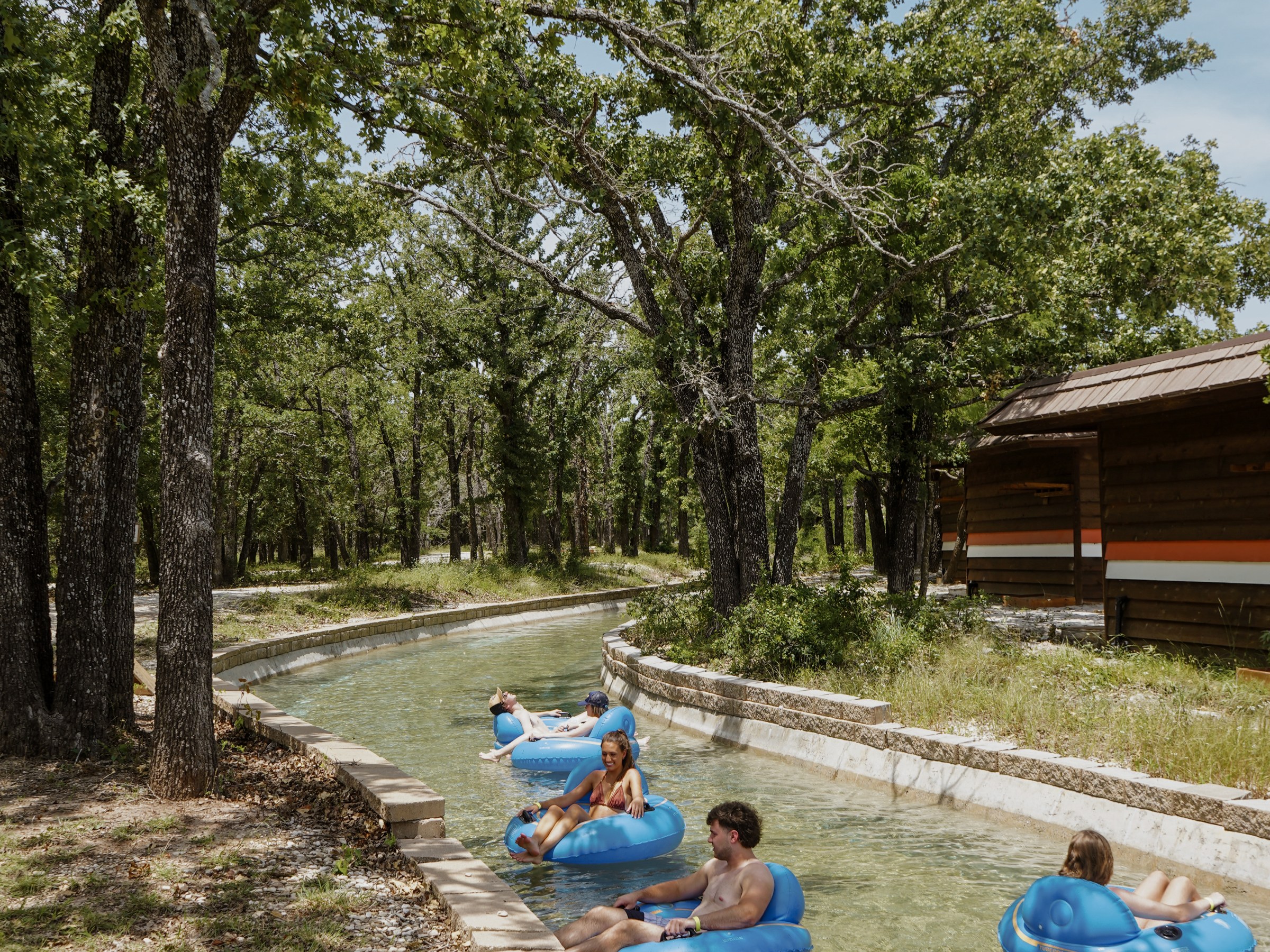 People float on blue tubes in a lazy river surrounded by trees and a wooden cabin.