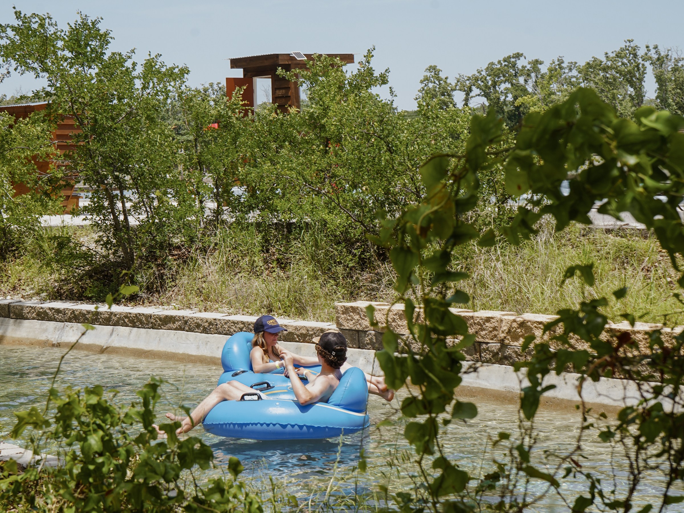 Two people relaxing on a blue inflatable in a lazy river surrounded by greenery.