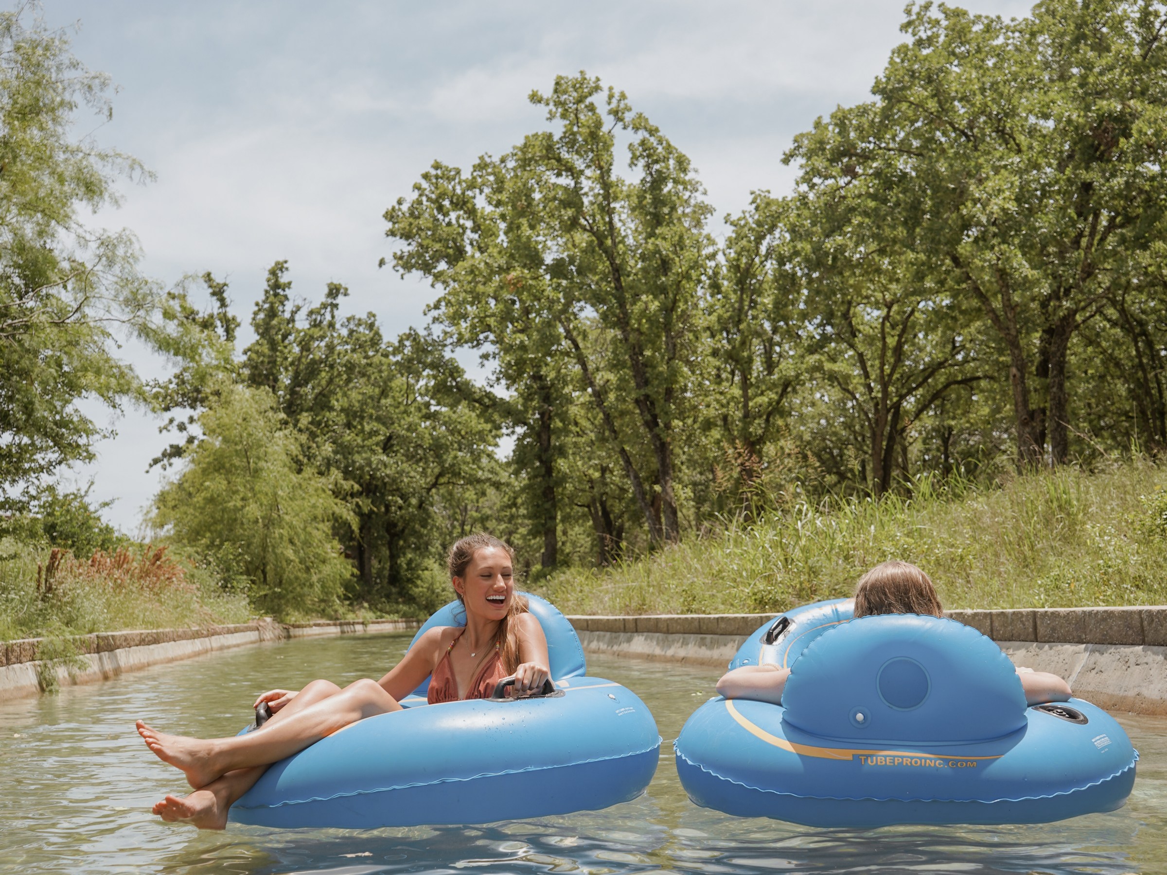 Two people relaxing on blue inflatables in a lazy river surrounded by trees.