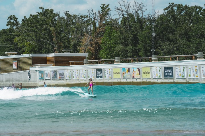 Person surfing on an artificial wave pool with trees and buildings in the background.