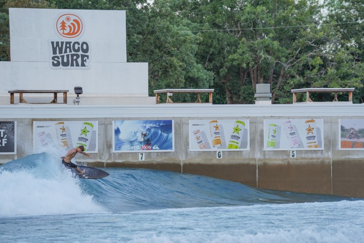 Surfer riding wave in outdoor wave pool at Waco Surf with trees and advertisements in background.
