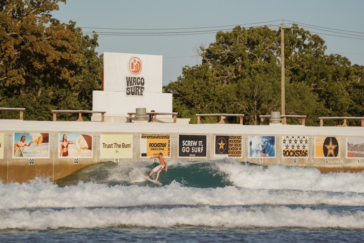 Person surfing in an artificial wave pool with advertising banners around.